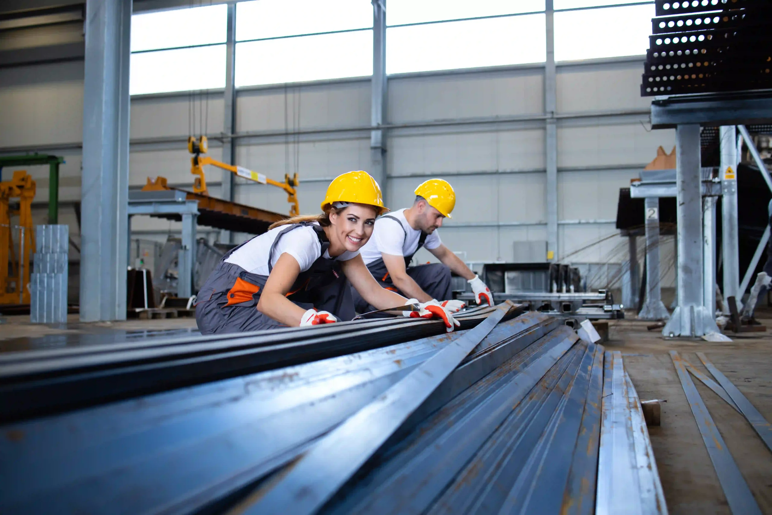 Two industrial workers wearing yellow safety helmets and gloves inspecting metal steel bars inside a factory hall.