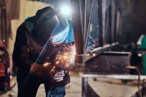 Seorang tukang las profesional mengenakan topeng pelindung wajah sedang mengelas kerangka besi hingga mengeluarkan Percikan api di bengkel pabrik.